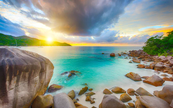 HD PC desktop wallpaper of a Seychelles beach at sunrise, showcasing the ocean, rocky shoreline, lush greenery, and a dramatic sky with clouds over the horizon.