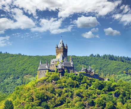 A view of Cochem Castle, a historic man-made castle nestled in a green forested hill in Germany under a blue sky with scattered clouds.