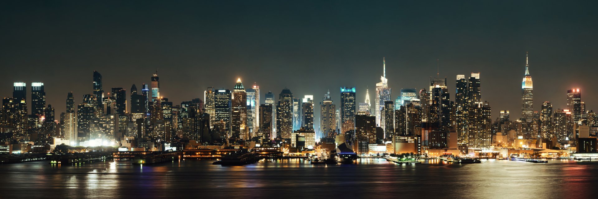Manhattan night skyline, man-made cluster of illuminated skyscrapers reflecting on the river, rendered as a 5K Ultra HD PC desktop wallpaper/background.
