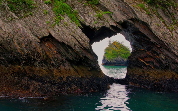 Natural rock arch frames a view of lush greenery and water in Alaska's Fjords National Park, showcasing rugged nature in this HD desktop wallpaper.