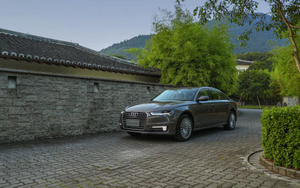 Silver Audi A6 parked on a stone driveway beside a brick wall, surrounded by greenery and trees, captured in 4K Ultra HD quality for a PC desktop wallpaper.