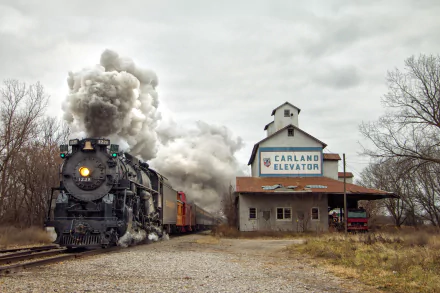 HD desktop wallpaper featuring a vintage steam locomotive emitting smoke, traveling past the Carland Elevator building on an overcast day.