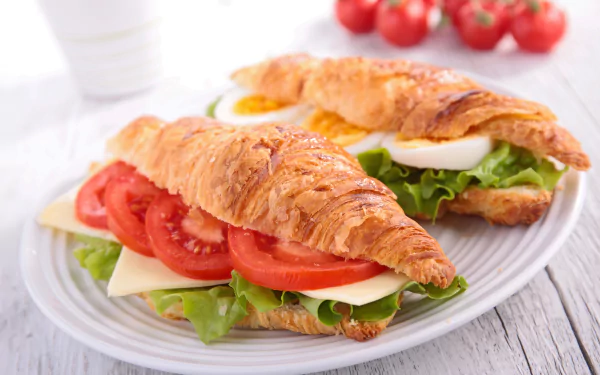 A 4K Ultra HD desktop wallpaper featuring fresh croissant sandwiches with lettuce, tomato, sliced egg, and cheese on a white plate, accompanied by cherry tomatoes.