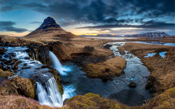 HD wallpaper of Kirkjufoss waterfall cascading beside the Kirkjufell mountain in Iceland, with a river winding through rugged natural terrain under a dramatic sky.