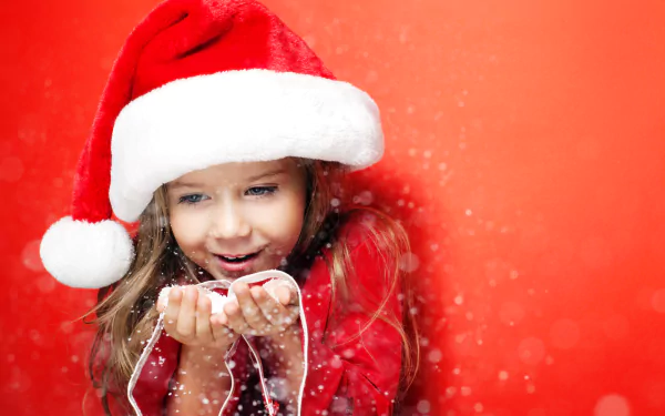 A young girl in a red Santa hat joyfully blows sparkling holiday snowflakes against a vibrant red background, captured in stunning 4K Ultra HD for a festive Christmas desktop wallpaper.
