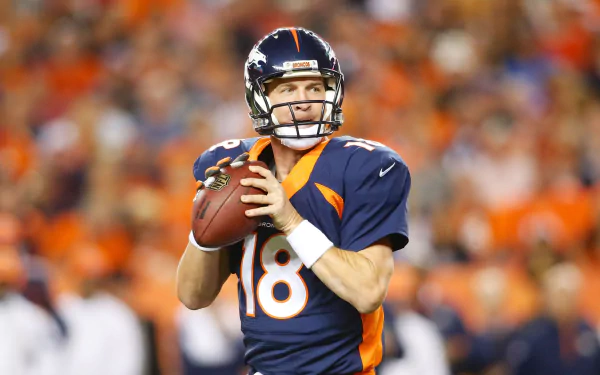 HD desktop wallpaper featuring a focused quarterback in a blue and orange uniform, poised to throw a football, with a vibrant crowd in the background.