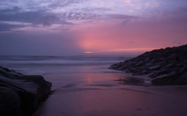 A serene beachscape with a pastel purple sunset over the horizon, reflecting on the calm sea and wet sand. Rocky formations frame the scene, creating a tranquil and picturesque nature HD wallpaper.