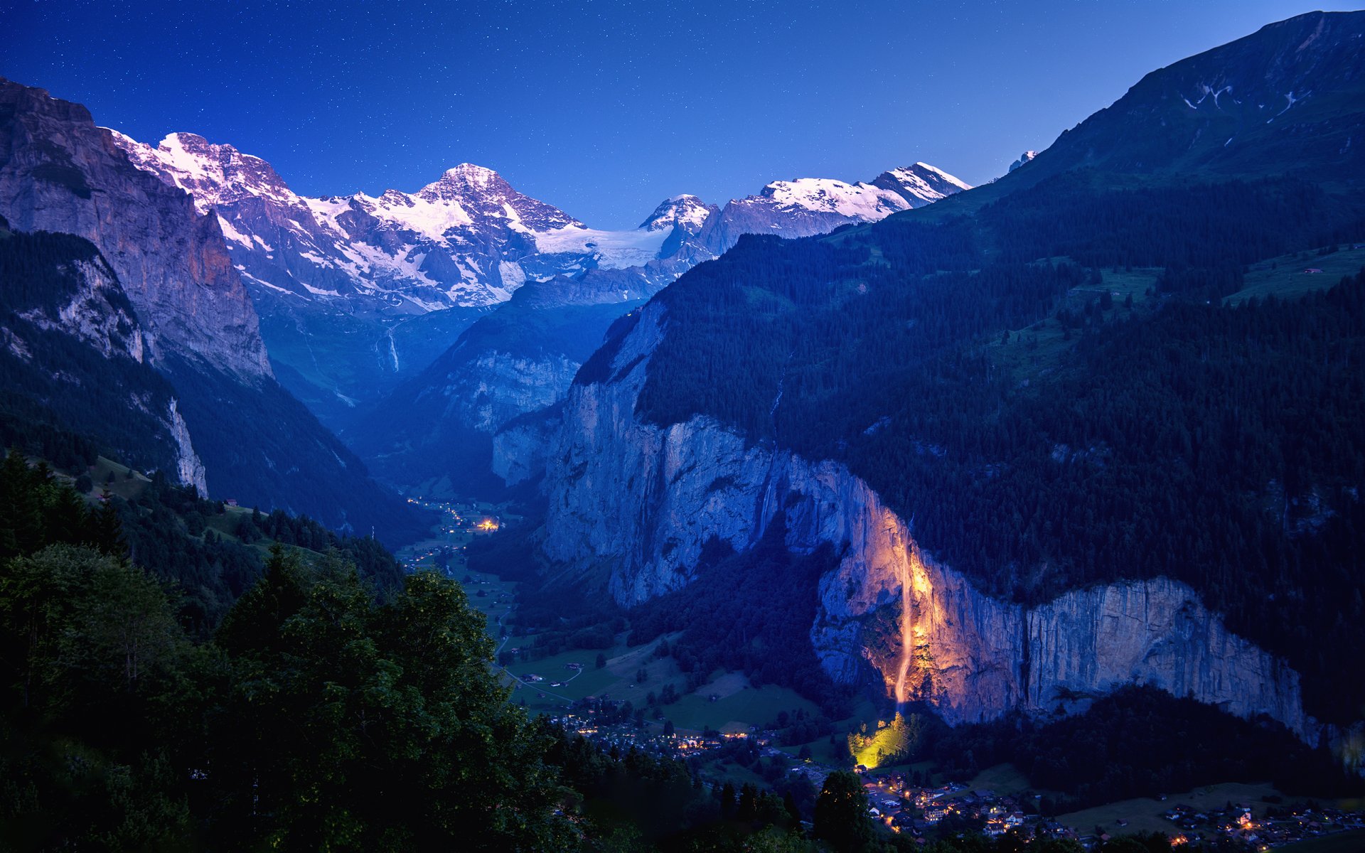 Nighttime landscape of a Swiss mountain valley illuminated by village lights, captured in high-definition photography for a PC desktop wallpaper background.