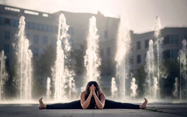A brunette woman practices yoga in a seated wide-leg pose in front of a large fountain, captured in an HD desktop wallpaper background.