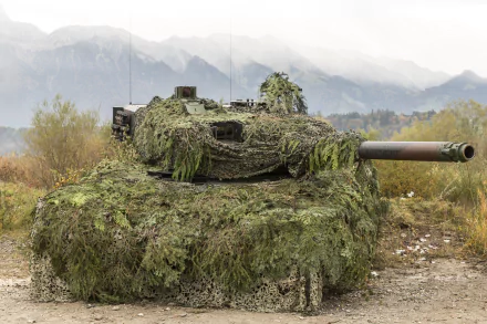Camouflaged Leopard 2 main battle tank draped in foliage netting on a dirt track, misty mountains behind — 5K Ultra HD PC desktop wallpaper.