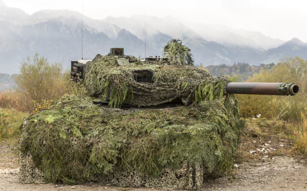 Camouflaged Leopard 2 main battle tank draped in foliage netting on a dirt track, misty mountains behind — 5K Ultra HD PC desktop wallpaper.