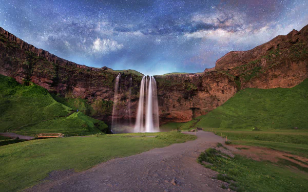 Stunning HD wallpaper of Seljalandsfoss waterfall in Iceland, framed by cliffs and a winding path, beneath a starry Milky Way sky showcasing nature's beauty.