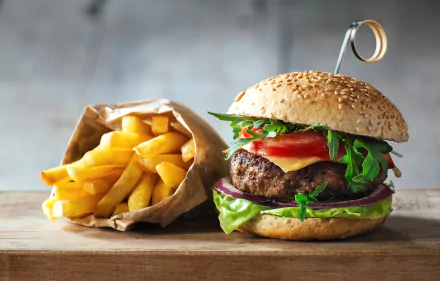 HD desktop wallpaper featuring a juicy burger with fresh greens and tomato, accompanied by a paper bag of golden french fries on a rustic wooden surface.