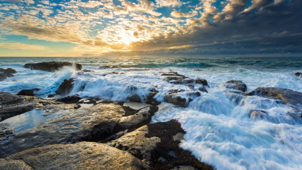 HD desktop wallpaper of a stunning seascape featuring ocean waves crashing over rocky shores at sunset, with a dramatic horizon and a sky full of scattered clouds.