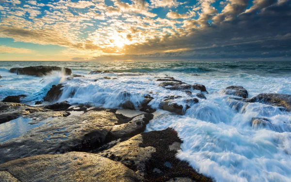 HD desktop wallpaper of a stunning seascape featuring ocean waves crashing over rocky shores at sunset, with a dramatic horizon and a sky full of scattered clouds.