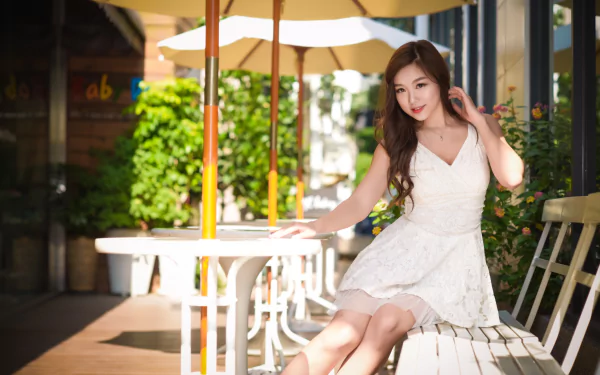 An Asian woman with brown eyes and brunette hair is seated, wearing a white dress. The background features blurred elements, giving depth of field. This photo is a high-definition desktop wallpaper.