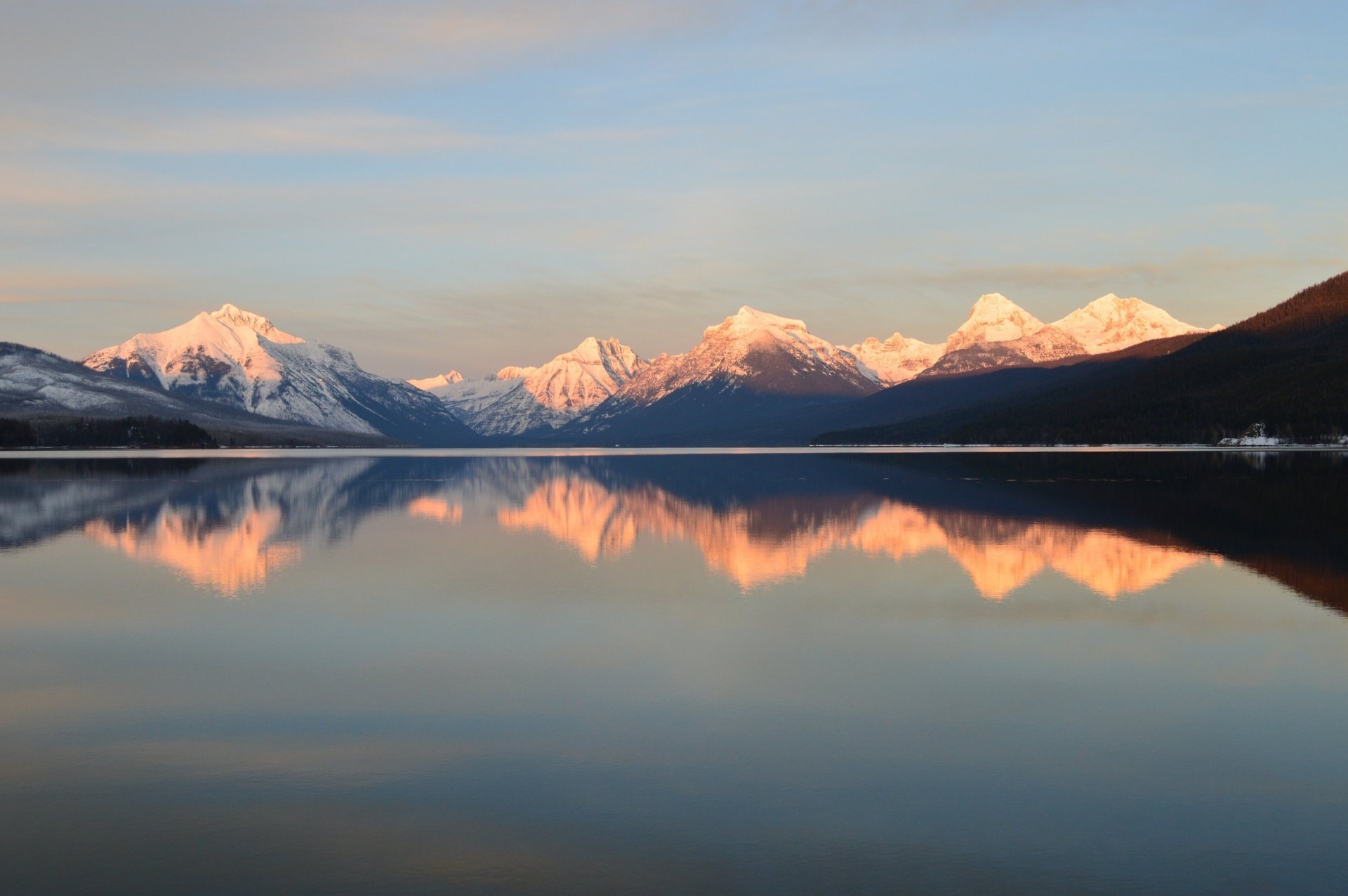 Snow-capped mountains of Glacier National Park reflect over the calm waters of Lake McDonald in Montana during winter, showcasing pristine wilderness and nature.