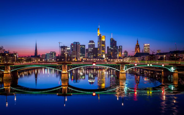 A stunning HD desktop wallpaper featuring the illuminated skyline of Frankfurt, Germany at night, with skyscrapers, bridges, and city lights reflecting on the calm river below.