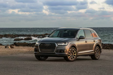 Silver Audi Q7 SUV parked by the ocean under a cloudy sky, captured in a high-definition desktop wallpaper showcasing the vehicle’s sleek design and rugged stance.