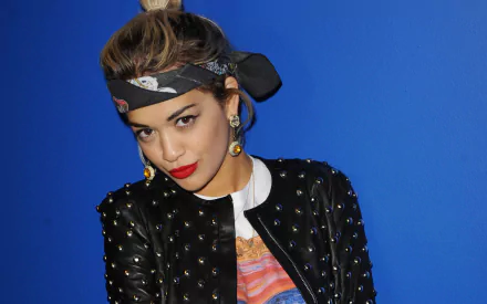 A HD desktop wallpaper featuring a brunette singer with brown eyes, wearing earrings, lipstick, and a studded jacket against a blue background. Her name is tagged as Rita Ora.