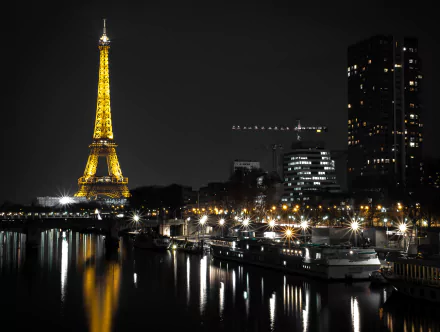 Nighttime view of the Eiffel Tower illuminated, reflecting on the Seine River in Paris with boats docked nearby and city lights enhancing the dark skyline.