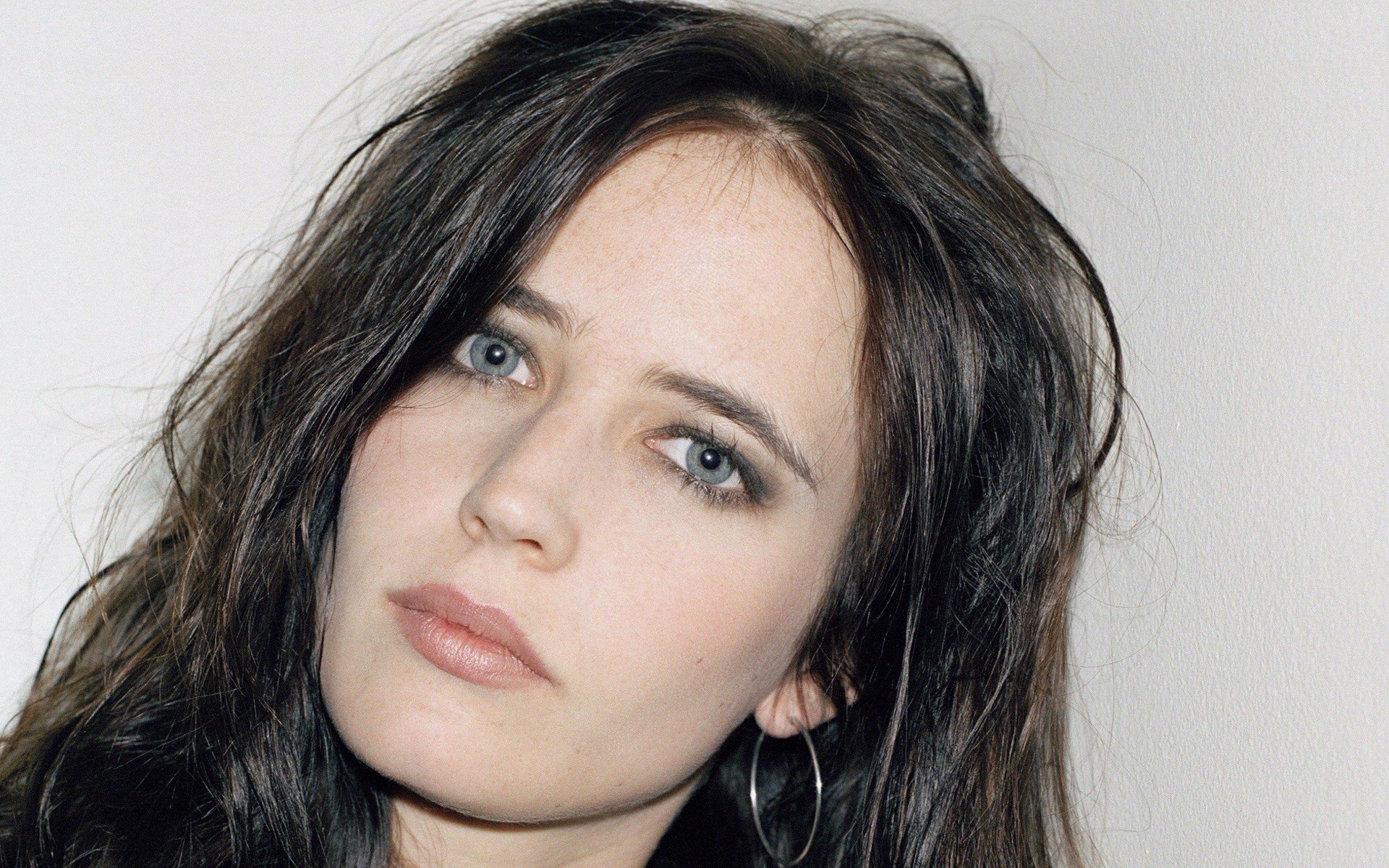 Close-up HD wallpaper of French actress Eva Green with striking blue eyes and brunette hair, featuring a soft, neutral expression against a plain background.