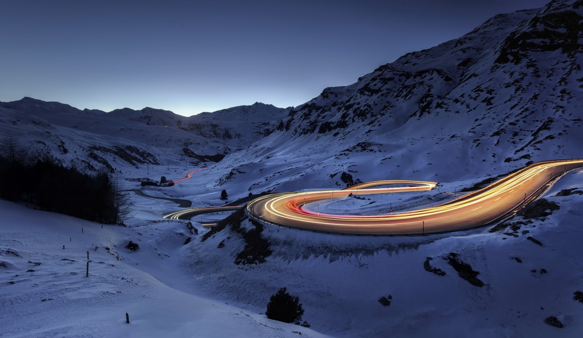 Nighttime winter landscape with snow-covered mountains and a winding road illuminated by streaks of light, captured in a stunning time-lapse photography HD wallpaper.