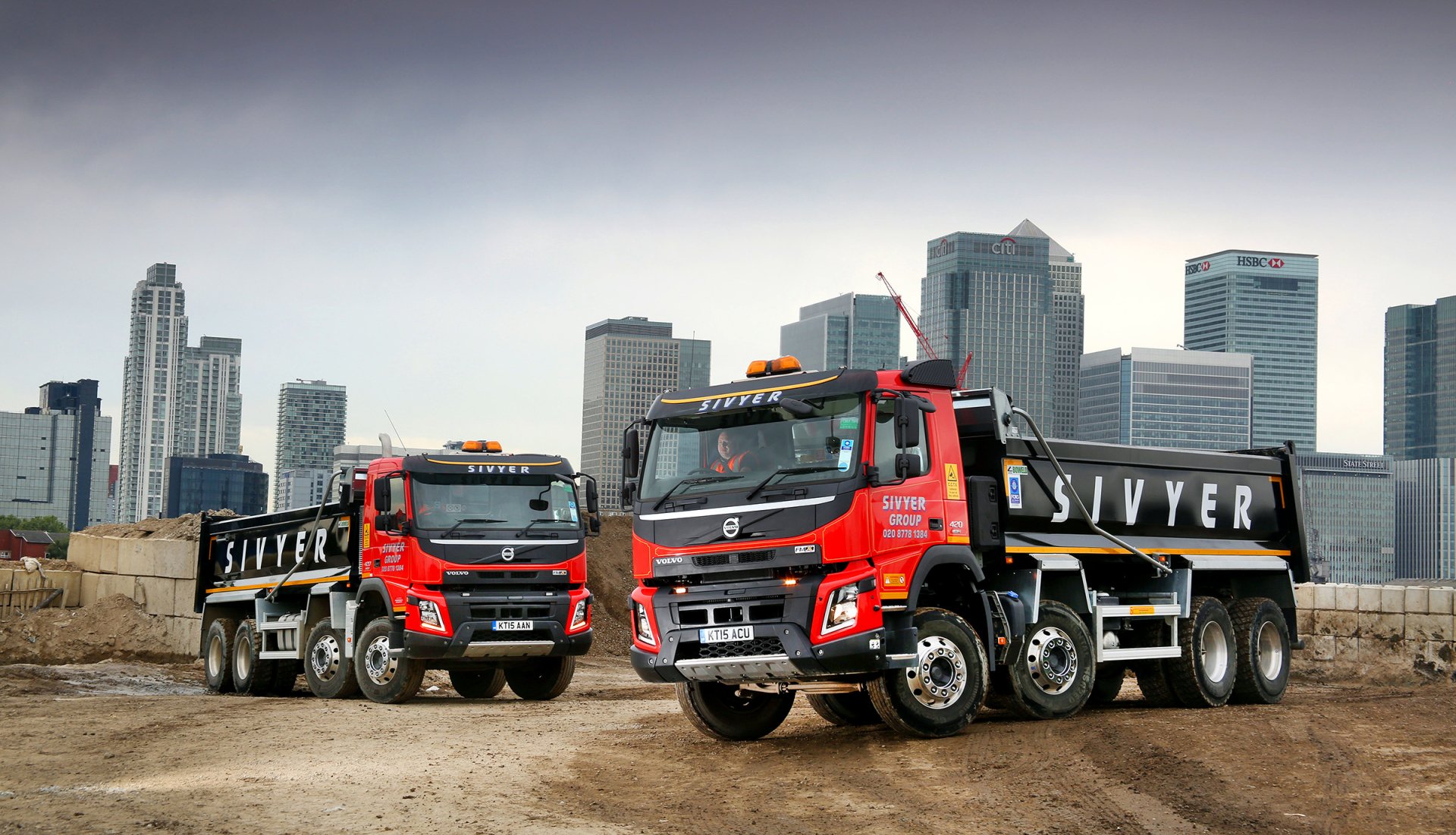 HD desktop wallpaper featuring two red Volvo dump trucks parked on a construction site with a city skyline in the background.