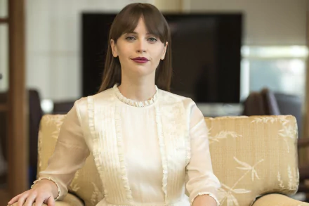 HD desktop wallpaper of a brunette celebrity actress seated on a beige sofa, wearing a white ruffled blouse, poised and facing the camera in a softly lit living-room background.