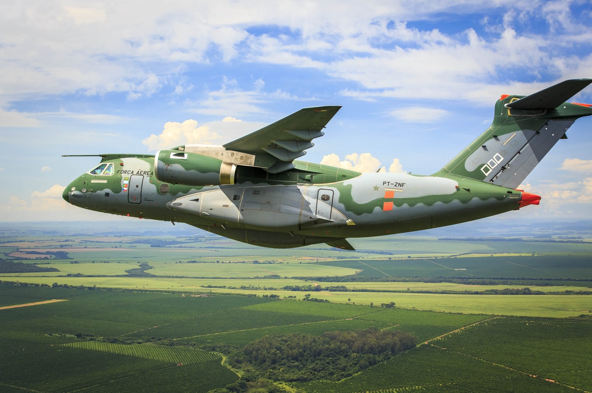 Embraer KC-390 military transport aircraft in flight over a green landscape under a partly cloudy sky, showcased as an HD PC desktop wallpaper.