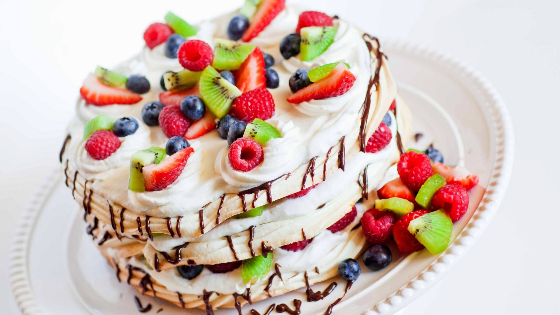 5K Ultra HD PC desktop wallpaper and background of a layered cream pastry cake topped with strawberries, raspberries, blueberries and kiwi slices, finished with chocolate drizzle.