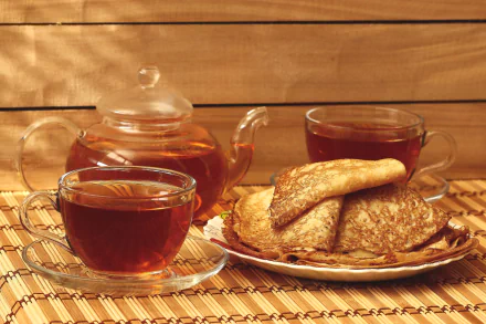 HD desktop wallpaper featuring a still life of a glass teapot, two cups of tea, and folded crêpes on a plate against a warm wooden background.