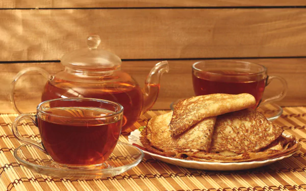HD desktop wallpaper featuring a still life of a glass teapot, two cups of tea, and folded crêpes on a plate against a warm wooden background.