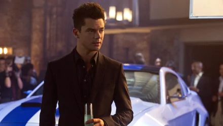 Dominic Cooper in a still from the movie Need For Speed, standing in front of a sports car, holding a drink. This HD desktop wallpaper captures a stylish scene from the film.