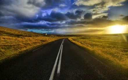 An HD desktop wallpaper featuring a man-made road stretching into the distance at sunset, with dramatic clouds and a setting sun casting a golden glow over surrounding fields.