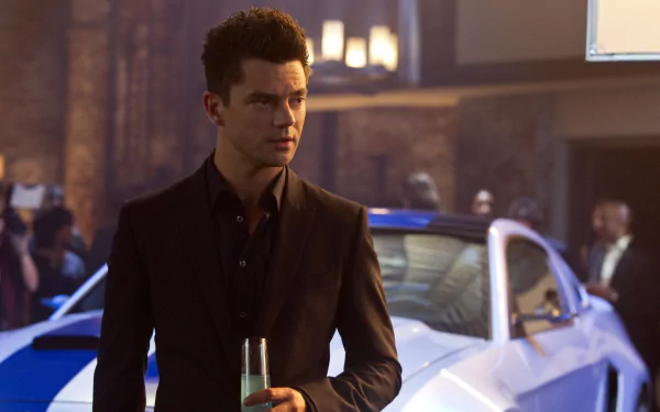 Dominic Cooper in a still from the movie Need For Speed, standing in front of a sports car, holding a drink. This HD desktop wallpaper captures a stylish scene from the film.