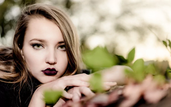 HD desktop wallpaper: close-up portrait of a woman model with green eyes and dark lipstick, face framed by soft bokeh and surrounding foliage.