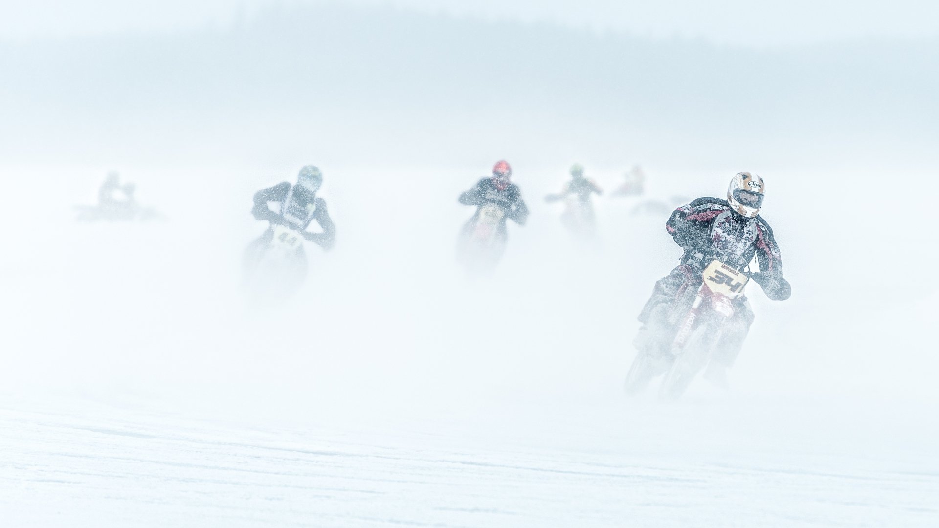 A high-definition wallpaper depicting a motocross race in snowy winter conditions, with riders emerging through a snowy mist, emphasizing the thrill and challenge of winter sports.
