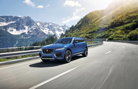 A blue Jaguar F-PACE SUV drives along a mountain road with snow-capped peaks and green hills under a bright sky, presented as a 4K Ultra HD desktop wallpaper.
