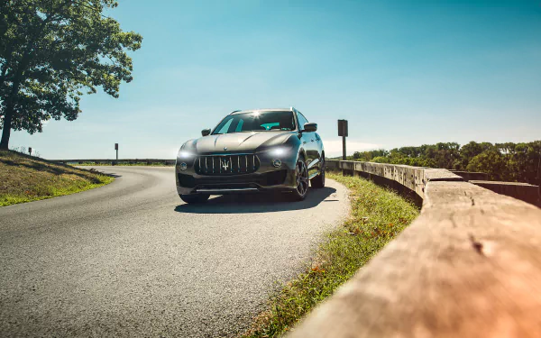 Silver Maserati Levante SUV on a winding road beside a wooden guardrail under a blue sky — HD PC desktop wallpaper and background.