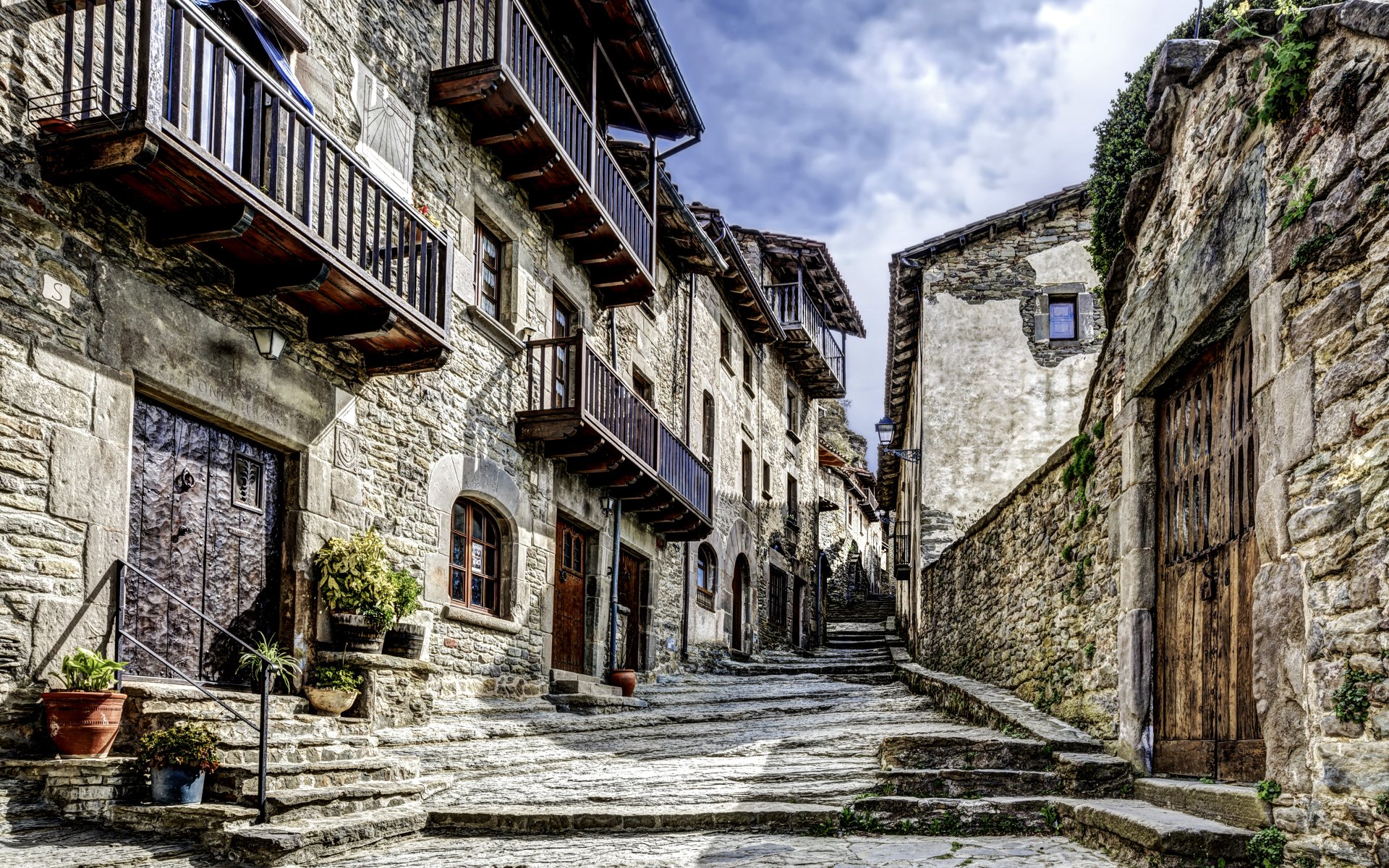 Stone-paved street in a Catalonian village, Spain, lined with rustic stone houses featuring wooden balconies under a cloudy sky, shown in 4K Ultra HD.