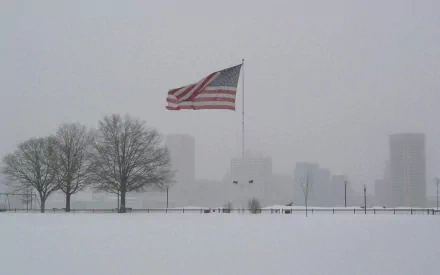A striking HD desktop wallpaper featuring the American flag waving against a snowy landscape, with bare trees and a faint city skyline in the background.