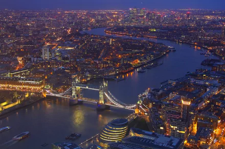 Aerial night view of London featuring the illuminated Tower Bridge over the Thames River and surrounding city buildings in the United Kingdom.
