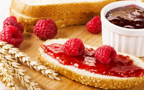 Close-up 4K Ultra HD wallpaper of raspberry jam spread on a slice of bread with fresh raspberries and wheat stalks, capturing a vibrant breakfast scene.