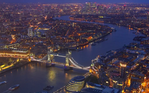 Aerial night view of London featuring the illuminated Tower Bridge over the Thames River and surrounding city buildings in the United Kingdom.