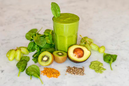 HD PC desktop wallpaper: avocado-kiwi green smoothie in a glass, surrounded by halved avocado, kiwi, spinach, grapes and assorted seeds on a marble background.