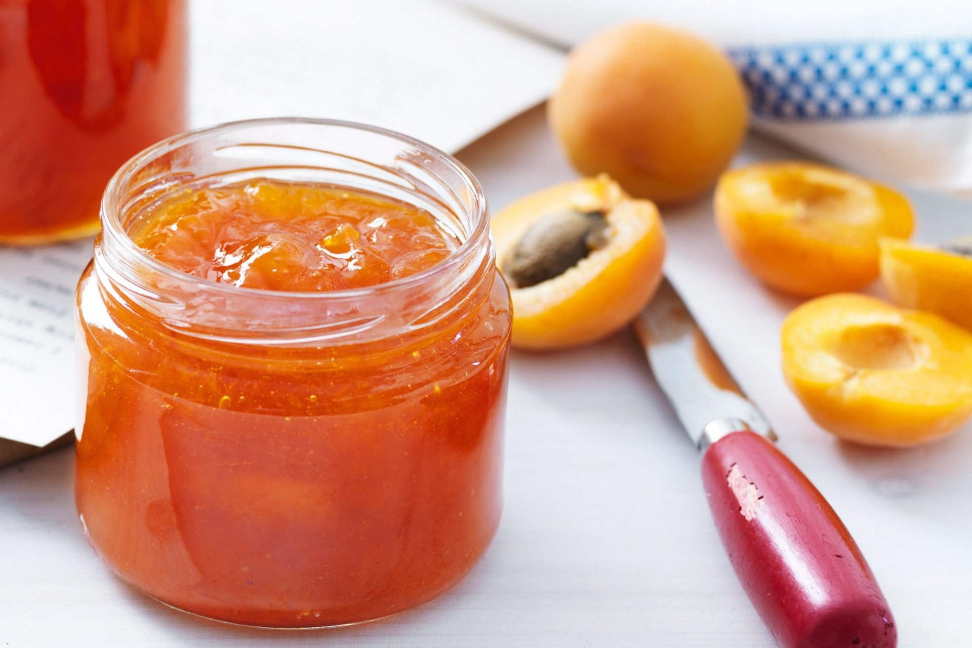 Close-up of a jar of apricot jam surrounded by fresh apricots and a knife on a white surface, captured as a vibrant HD PC desktop wallpaper and background.