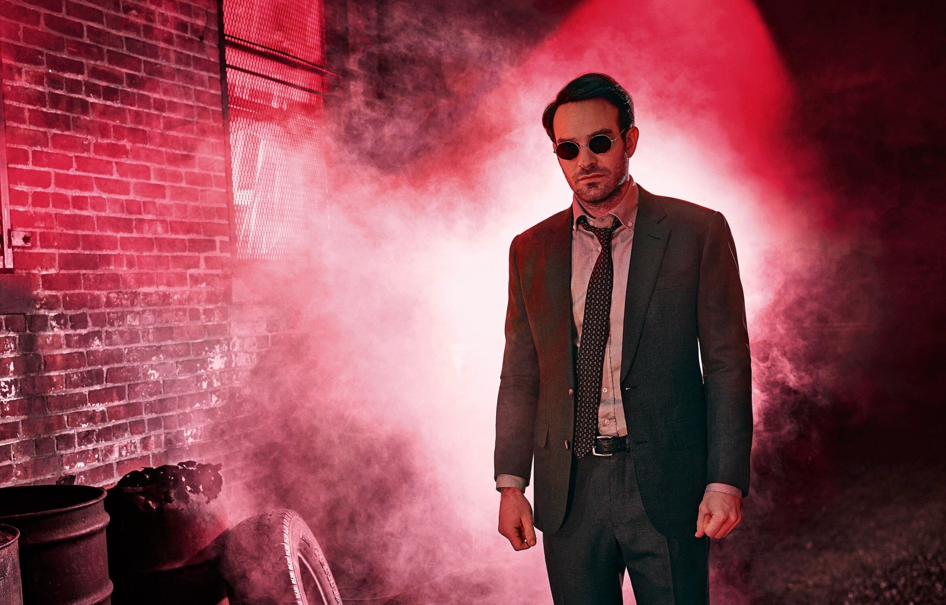 Charlie Cox as Daredevil stands in a smoky, red-lit alley, dressed in a suit and sunglasses, from the TV shows Daredevil and The Defenders.