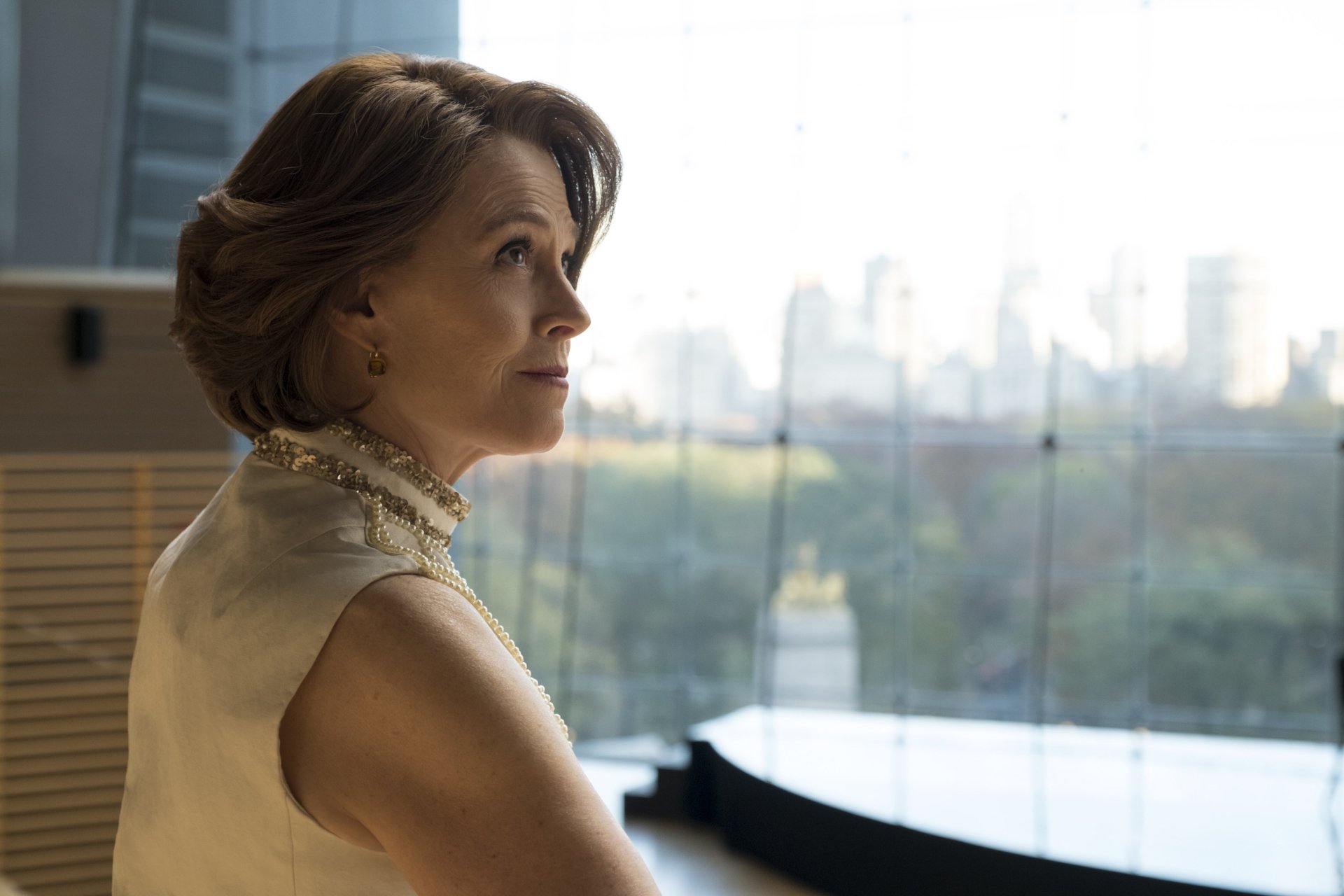 HD desktop wallpaper featuring Alexandra (Marvel) from the TV Show The Defenders, portrayed by Sigourney Weaver, against a backdrop with cityscape views through large glass windows.