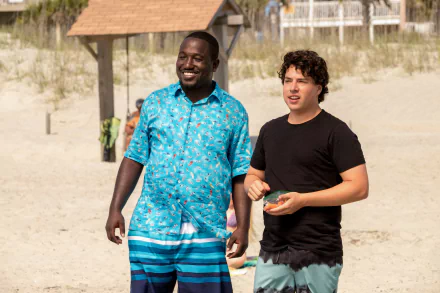 A HD wallpaper featuring Jon Bass and Hannibal Buress from the movie Baywatch. They are standing on a sunny beach with smiles, dressed in casual beachwear.